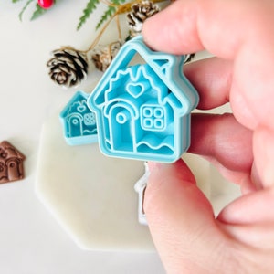 May include: A set of two blue plastic gingerbread house cookie cutters. The larger cutter has a heart and window details. The smaller cutter is a simple gingerbread house shape.