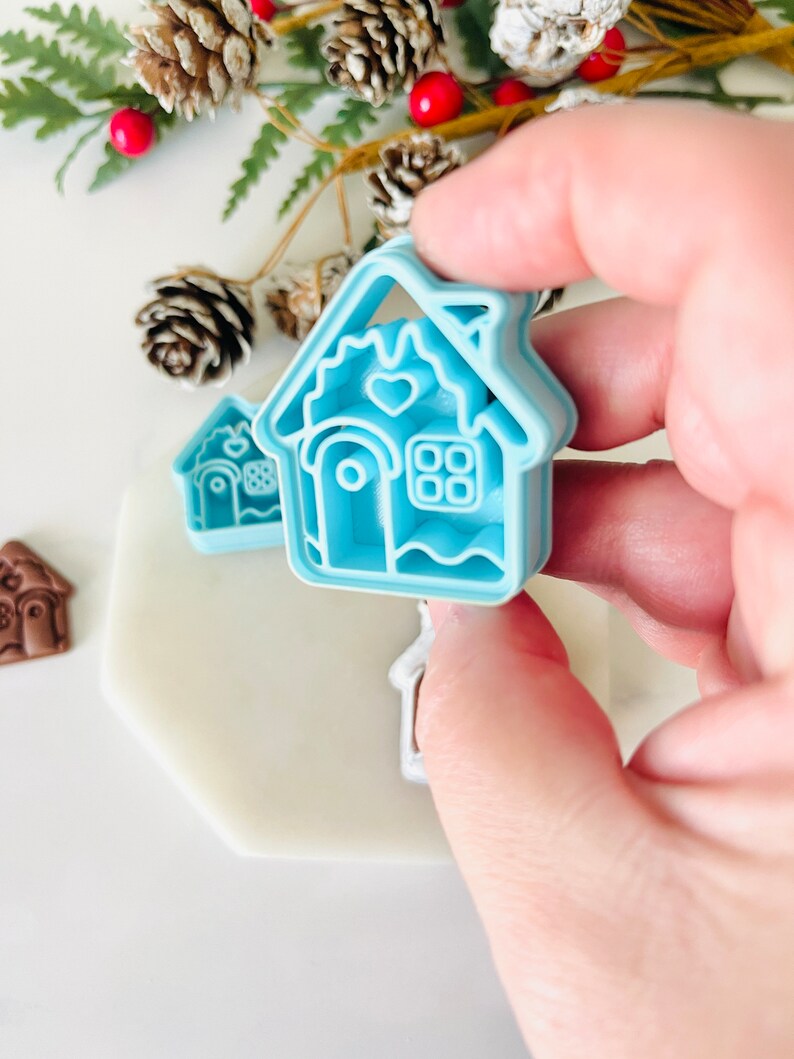 May include: A set of two blue plastic gingerbread house cookie cutters. The larger cutter has a heart and window details. The smaller cutter is a simple gingerbread house shape.