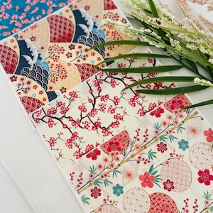 May include: A selection of Japanese-style patterned papers. Designs feature cherry blossoms, waves, and geometric shapes in red, blue, and cream. The papers are arranged in a layered display, showcasing the variety of patterns.
