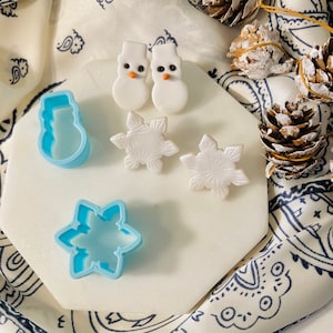 May include: A white ceramic tray with two white snowman-shaped ornaments, two white snowflake-shaped ornaments, and two blue plastic cookie cutters shaped like a snowman and a snowflake.