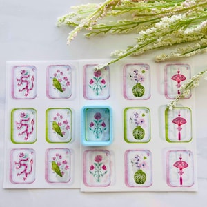 May include: A sheet of decorative stickers featuring watercolor illustrations of dragons, birds, flowers, and decorative objects. The stickers have a white background with pink and green borders. A small, light blue container is also present.