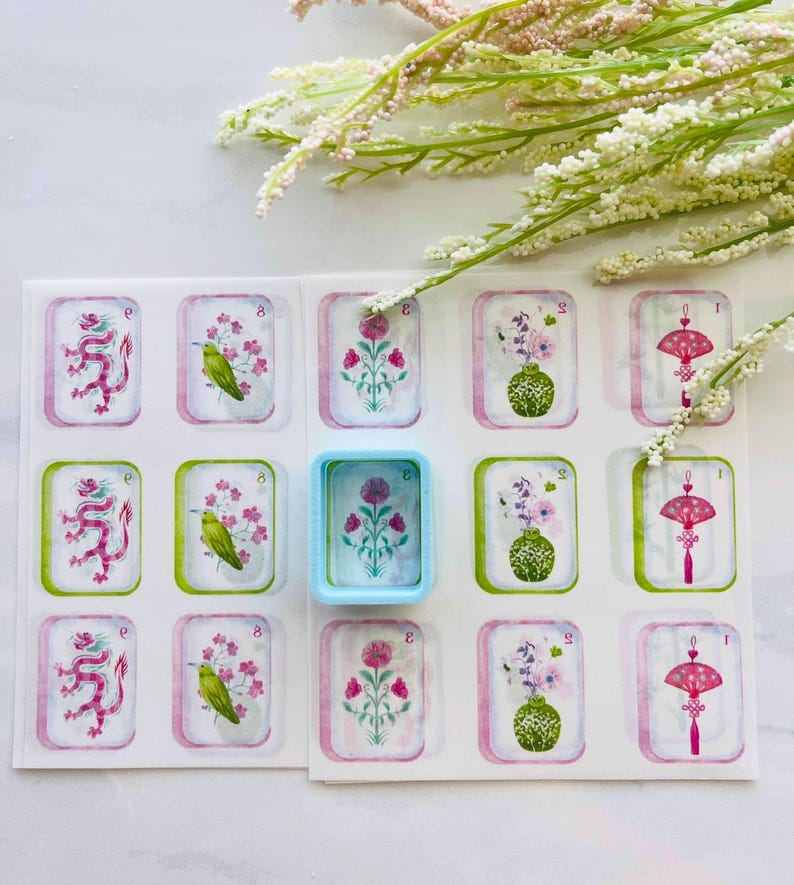 May include: A sheet of decorative stickers featuring watercolor illustrations of dragons, birds, flowers, and decorative objects. The stickers have a white background with pink and green borders. A small, light blue container is also present.