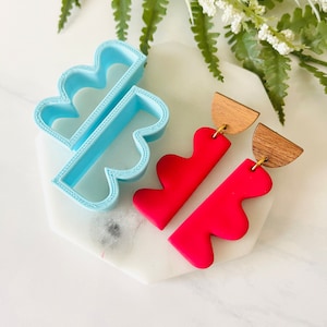 Könnte beinhalten: Ein Paar leuchtend rote Ohrringe aus Polymer Clay mit einer wellenförmigen, organischen Form. Die Ohrringe haben ein oberes Teil aus Holz und goldfarbene Haken.