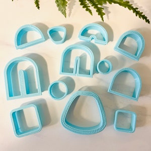 May include: A set of 11 blue plastic cookie cutters in various shapes, including arches, circles, squares, and a teardrop shape. The cutters have a decorative border around the edges.