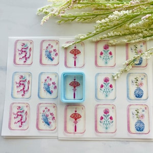 May include: A sheet of decorative stickers featuring various designs, including a red dragon, a blue bird with pink flowers, and a red fan with a tassel. A light blue container holds a sticker with a red fan design. The stickers have a watercolor effect.