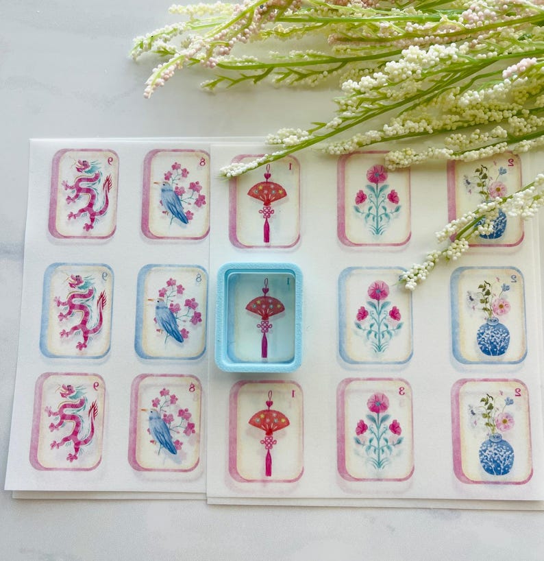 May include: A sheet of decorative stickers featuring various designs, including a red dragon, a blue bird with pink flowers, and a red fan with a tassel. A light blue container holds a sticker with a red fan design. The stickers have a watercolor effect.