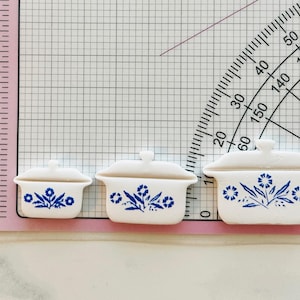 May include: Three miniature white ceramic casserole dishes with lids, adorned with blue floral patterns. The dishes are arranged in a row, varying in size, with the largest measuring approximately 1.5 inches in length.