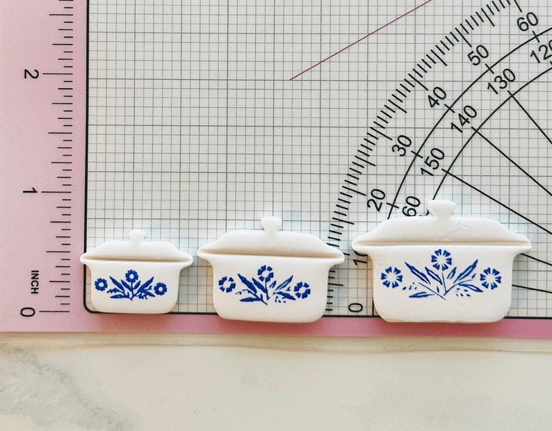 May include: Three miniature white ceramic casserole dishes with lids, adorned with blue floral patterns. The dishes are arranged in a row, varying in size, with the largest measuring approximately 1.5 inches in length.