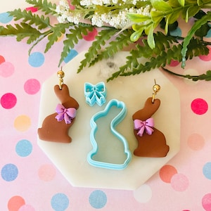 May include: Two brown bunny shaped earrings with purple bows, a blue bunny shaped cookie cutter, and a blue bow cookie cutter on a white surface with pink and blue polka dots.