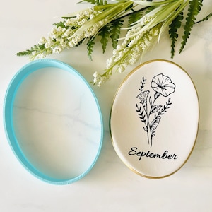 Pode incluir: Um prato de cerâmica branco com uma borda dourada apresenta um desenho de linha preta de flores e a palavra "Setembro". O prato está ao lado de um anel oval azul claro.