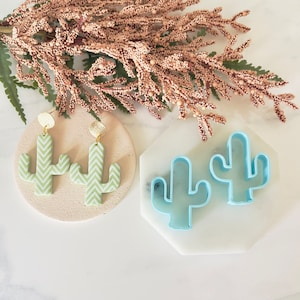 May include: Two green and white chevron patterned cactus earrings with gold accents and two blue plastic cactus cookie cutters on a white surface.