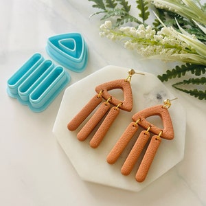 May include: A pair of brown clay earrings with a geometric triangle shape and dangling rectangular shapes. The earrings have gold accents.