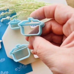 May include: A light blue plastic cookie cutter in the shape of a cooking pot is held in a person's hand. Two additional cutters are visible in the background. The cutters are on a white and blue patterned surface.