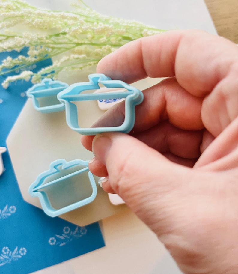 May include: A light blue plastic cookie cutter in the shape of a cooking pot is held in a person's hand. Two additional cutters are visible in the background. The cutters are on a white and blue patterned surface.