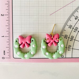 May include: A pair of green and white polka dot wreath earrings with pink bows. The earrings are made of polymer clay and have gold hooks.