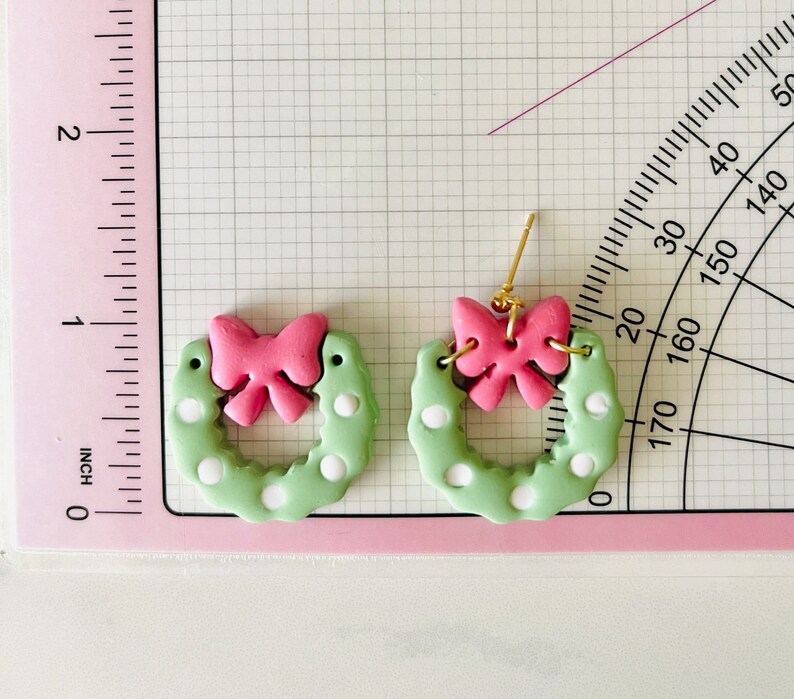 May include: A pair of green and white polka dot wreath earrings with pink bows. The earrings are made of polymer clay and have gold hooks.