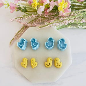 May include: A set of six blue and yellow plastic cookie cutters shaped like baby chicks. The cutters are arranged on a white surface.