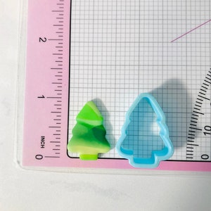 May include: A green and yellow Christmas tree cookie cutter and a blue plastic Christmas tree cookie cutter on a white grid background with a pink ruler.