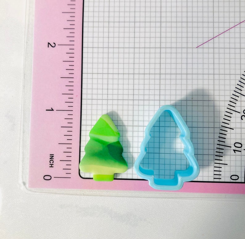 May include: A green and yellow Christmas tree cookie cutter and a blue plastic Christmas tree cookie cutter on a white grid background with a pink ruler.