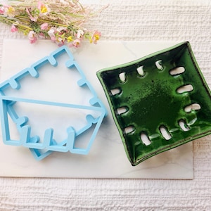 May include: A light blue plastic berry basket cookie cutter positioned next to a square green ceramic berry basket with a glossy finish and multiple drainage holes along the sides and base, both resting on a white marble surface.