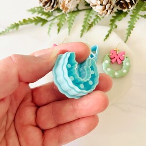 May include: A pair of handmade polymer clay earrings in the shape of a Christmas wreath. One earring is light blue with white polka dots, and the other is green with white polka dots and a pink bow.