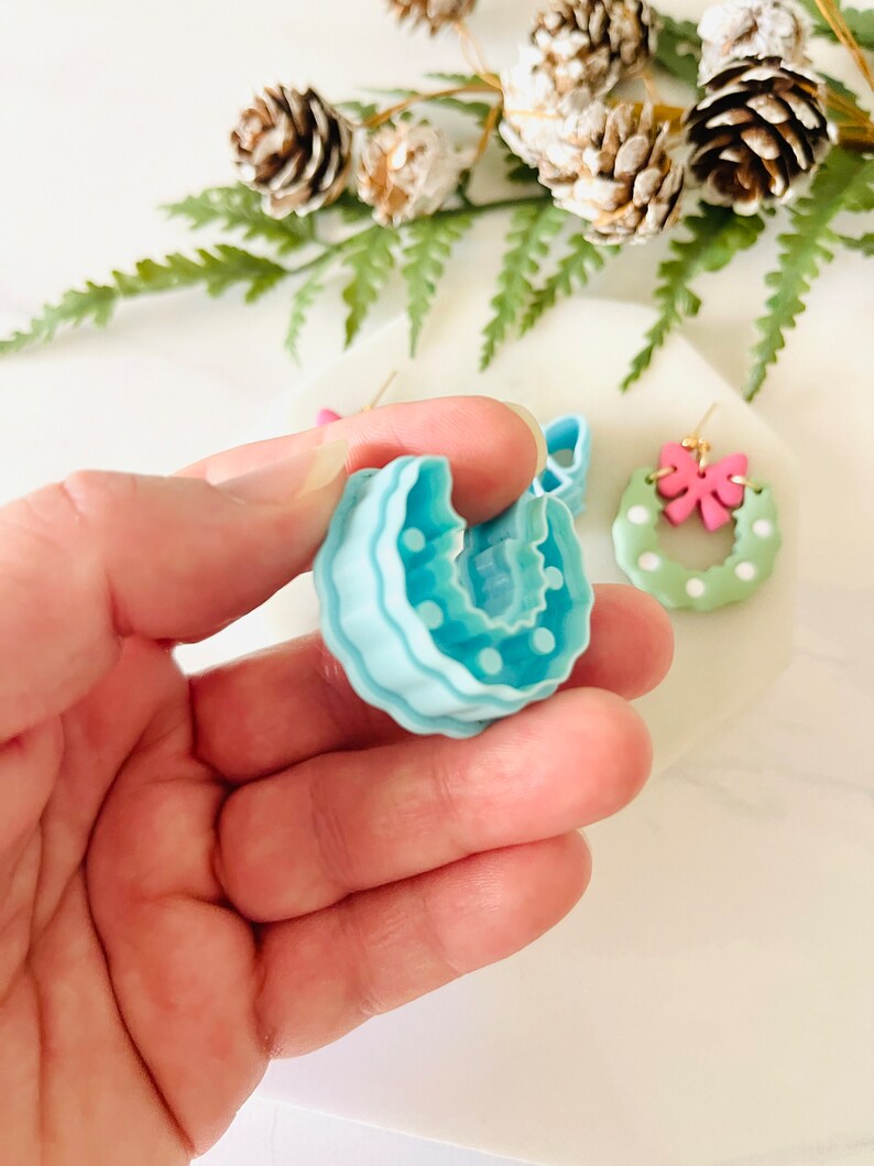 May include: A pair of handmade polymer clay earrings in the shape of a Christmas wreath. One earring is light blue with white polka dots, and the other is green with white polka dots and a pink bow.