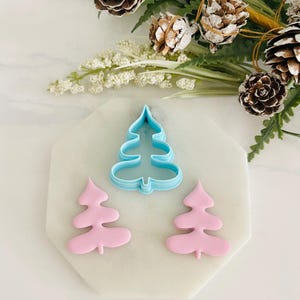 May include: A set of three cookie cutters in the shape of Christmas trees. The cutters are made of blue and pink plastic. The larger cutter is blue and the two smaller cutters are pink.
