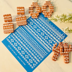 May include: A set of terracotta earrings and cactus ornaments with white geometric patterns. The earrings are in the shape of a square and the cactus ornaments are in the shape of a traditional cactus. The ornaments are on a blue background with a white stencil of geometric patterns. The stencil reads "SO CAL CUTTER CO. AZTEC PATTERN".