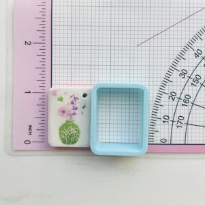 May include: A small, rectangular ceramic pendant with a floral design featuring a green vase and purple flowers. A light blue square-shaped object is next to the pendant. The pendant is approximately 1 inch in length.