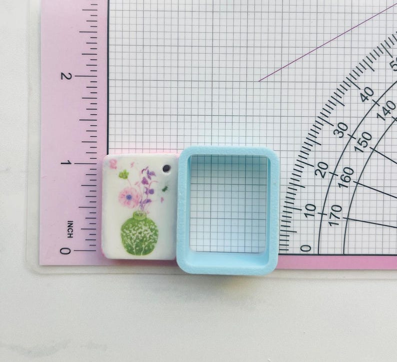 May include: A small, rectangular ceramic pendant with a floral design featuring a green vase and purple flowers. A light blue square-shaped object is next to the pendant. The pendant is approximately 1 inch in length.