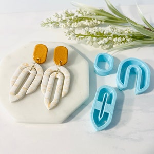 May include: A set of three blue polymer clay cutters for making earrings. The cutters are shaped like an oval, a half-circle, and a long, thin rectangle. The cutters are on a white surface next to a pair of earrings made with white polymer clay and gold accents.