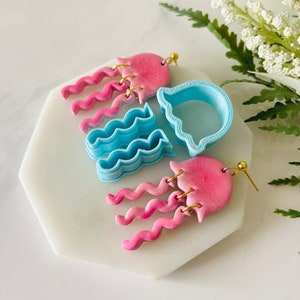 Pode incluir: Um par de brincos de argila polimérica rosa e azul com um design de água-viva. Os brincos são feitos à mão e têm um tema caprichoso inspirado no oceano.