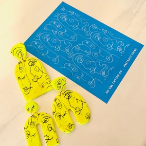 Può includere: Un paio di orecchini gialli a forma di arco con un design di viso astratto nero. Gli orecchini sono in argilla polimerica. Gli orecchini sono mostrati su uno sfondo blu con il testo "SO CAL CUTTER CO. ABSTRACT FACES".