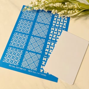 Puede incluir: Una hoja de plantilla azul con patrones de azulejos geom&eacute;tricos blancos. La hoja tiene el texto "SO CAL CUTTER CO. GEO TILE PATTERNS" impreso en ella. Una tarjeta blanca en blanco se coloca encima de la hoja de plantilla.