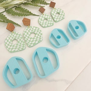 Könnte beinhalten: Ein Set aus vier Polymer Clay Ohrringen in einem grün-weiß karierten Muster mit einem Loch in der Mitte. Die Ohrringe sind mit Holzstiften gepaart. Es gibt auch zwei blaue Polymer Clay Formen in Form eines abgerundeten Rechtecks mit einem Loch in der Mitte.
