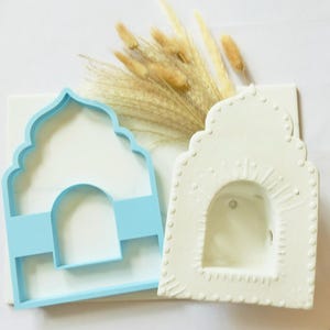 Puede incluir: Un cortador de galletas de plástico azul claro con forma de arco junto a una pieza decorativa de arcilla o cerámica blanca con forma de arco, que presenta un borde de puntos en relieve y un recorte central, ambos con una silueta arquitectónica.