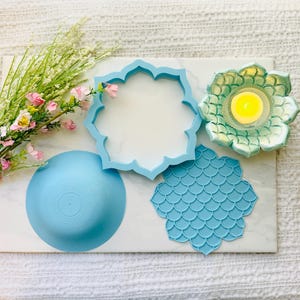 Puede incluir: Un juego de moldes de silicona azul claro para manualidades, que incluye un molde de cuenco circular, un marco con forma de loto y un inserto texturizado de pétalos de loto, junto a un portavelas de loto verde claro con una vela amarilla.