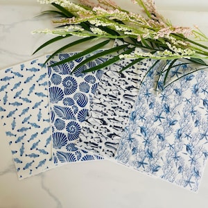 May include: Four white fabric napkins with blue marine patterns. Designs include fish, seashells, and coral. The napkins are arranged on a white surface with green and pink floral accents.