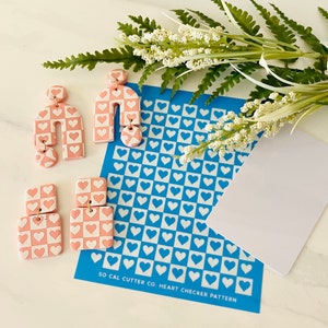 May include: Pink and white heart checkerboard pattern polymer clay earrings with gold hoops. The earrings are displayed on a blue sheet with a white heart checkerboard pattern. The sheet has the text "SO CAL CUTTER CO: HEART CHECKER PATTERN".