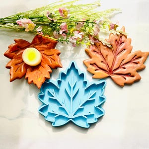 May include: Two ceramic maple leaf dishes in shades of orange and brown, one holding a small round yellow candle, alongside a light blue plastic leaf-shaped mold. A cluster of small pink and white flowers rests above the items.