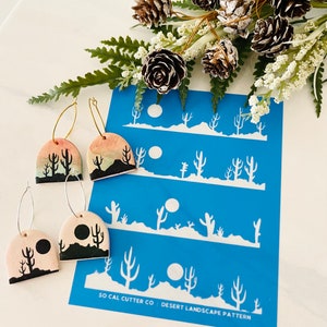 May include: A pair of ceramic earrings with a desert landscape design. The earrings are painted with a sunset gradient and feature a silhouette of cacti and a moon. The earrings are shown next to a blue stencil with the text "SO CAL CUTTER CO. : DESERT LANDSCAPE PATTERN".