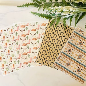 May include: Four sheets of decorative paper with different western-themed patterns. One sheet features pink cowboy boots, cacti, and skulls. Another sheet has a pattern of brown horseshoes. The third sheet has a pattern of brown and blue geometric shapes. The final sheet has a pattern of brown and blue geometric shapes.