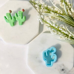 Puede incluir: Dos adornos de cactus de arcilla verde con acentos rojos y un cortador de galletas de cactus de plástico azul claro sobre una superficie de mármol blanco.