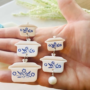 May include: White and blue casserole dish earrings with a floral design. The earrings are made of a lightweight material and feature a dangling pearl. The earrings are a unique and fun accessory.