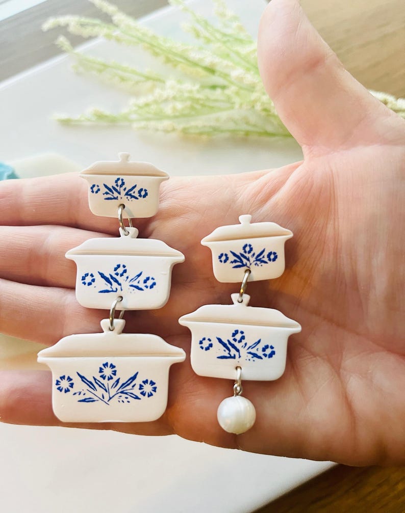 May include: White and blue casserole dish earrings with a floral design. The earrings are made of a lightweight material and feature a dangling pearl. The earrings are a unique and fun accessory.