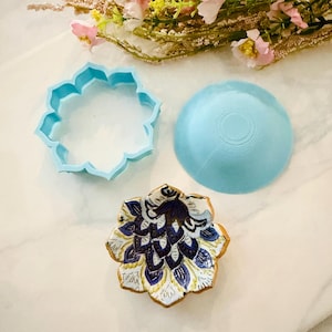 May include: A light blue flower-shaped cookie cutter and a matching blue bowl are next to a decorated cookie. The cookie is in the shape of a flower with blue, white, and gold details. The items are on a white surface.