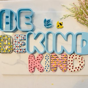 Puede incluir: Una composición decorativa que deletrea "BE KIND" con moldes de letras azules y letras coloridas con patrones. Las letras están sobre una superficie blanca similar al mármol, con pequeños adornos florales y con forma de insectos.