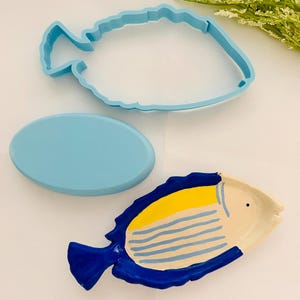 May include: A light blue fish-shaped cookie cutter and a matching oval-shaped object are next to a hand-painted ceramic fish dish. The dish is white with yellow and blue details, and blue stripes.