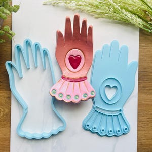 以下が含まれることがあります： 3つの手型クッキーカッター。1つは水色、もう1つはピンクのスカートと中央にハートが付いた手、3つ目は中央にハートが付いた手型クッキーカッターです。