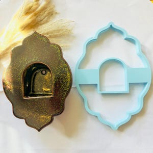May include: A dark brown ceramic wall-mounted niche with a speckled gold finish and a central arched opening, positioned next to a light blue plastic cookie cutter that mirrors the same quatrefoil shape and arched cutout design.